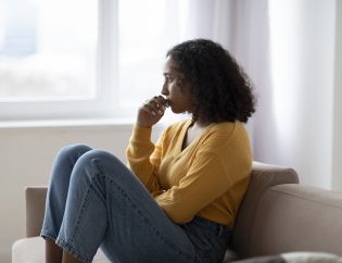 A young black woman sitting on her couch looking sad. She is wearing a yellow shirt and blue jeans.