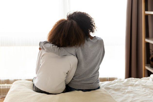 Two women sitting on a bed. They both are facing away from the camera. One has their arm around the other. Both of their heads are leading on one another's shoulders.