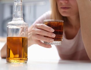 a woman holding a whiskey glass filled to the brim. The glass whiskey bottle is sitting next to her. She looks depressed and has her arm and hand on her head.