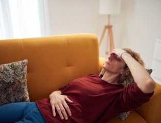 An older woman laying on a couch in pain with her hand over her eyes and one hand over her stomach.