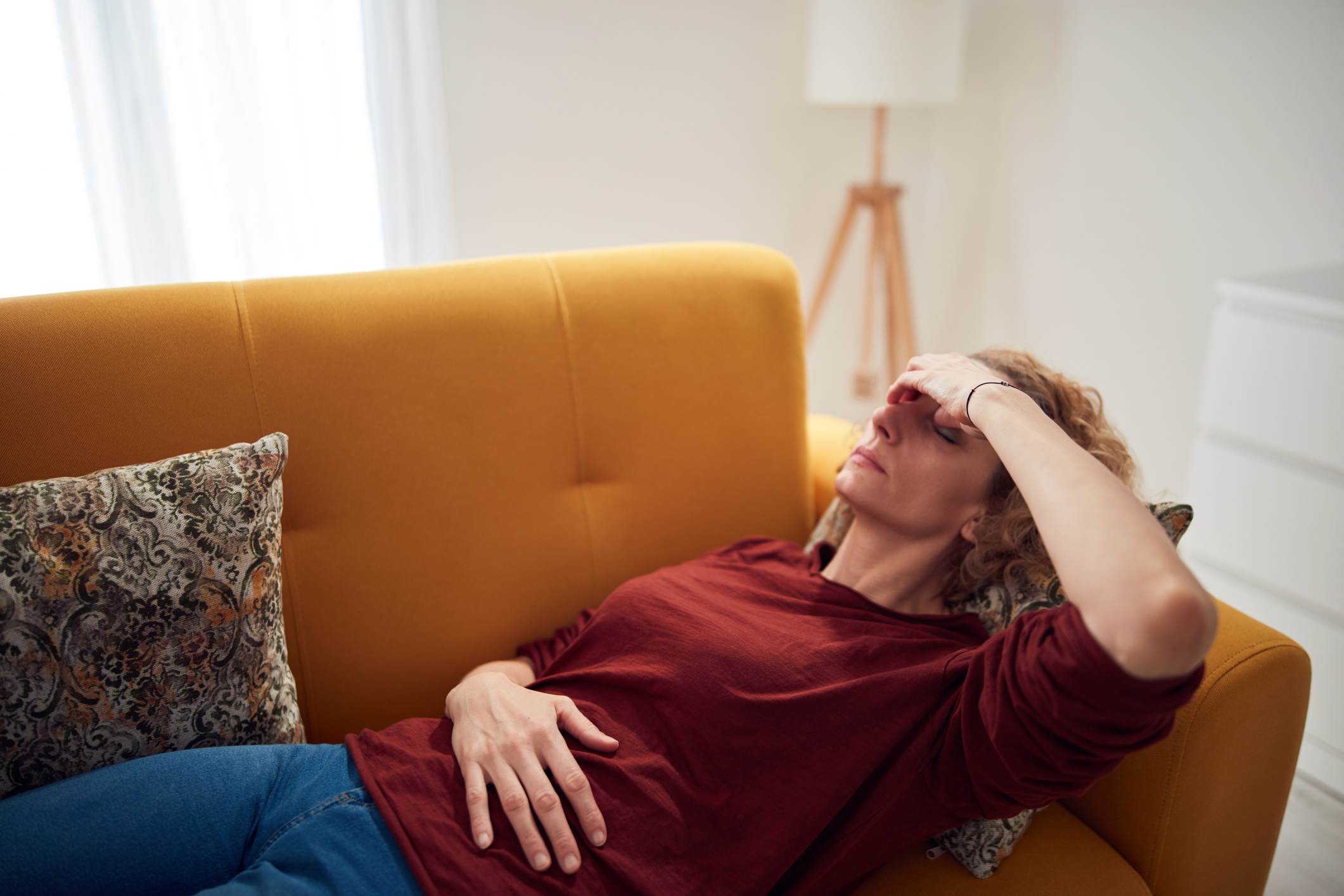 An older woman laying on a couch in pain with her hand over her eyes and one hand over her stomach.