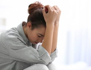 A young woman experiencing depression. She has her arms on her knees and her hands are resting on her head. She is looking down to the floor sadly.