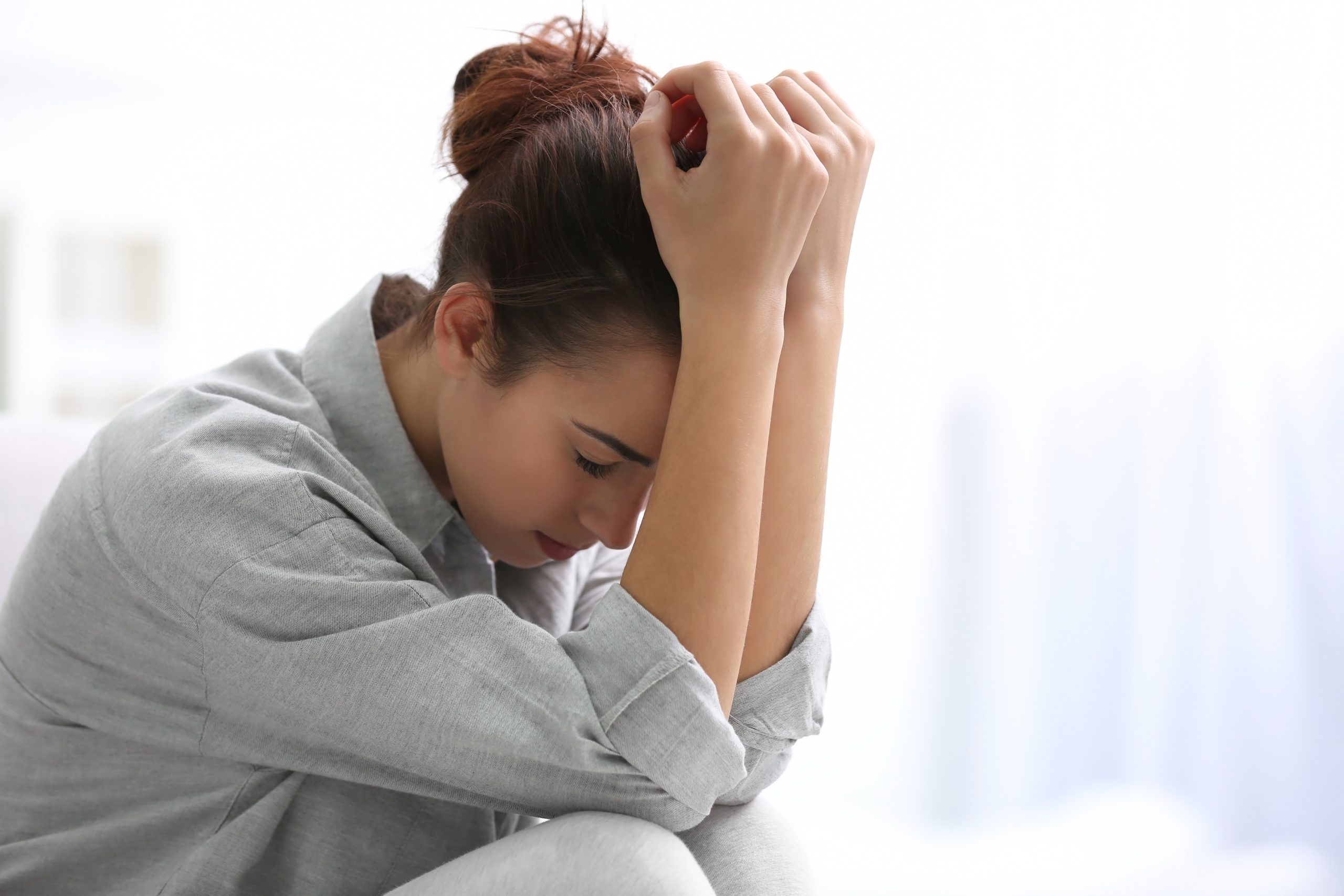 A young woman experiencing depression. She has her arms on her knees and her hands are resting on her head. She is looking down to the floor sadly.