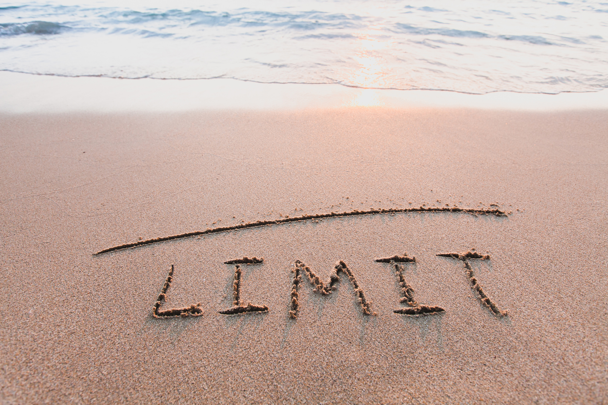 The word "limit" written in the sand with a line above it, representing how to set healthy boundaries. The ocean waves are crashing onto the sand in the distance.