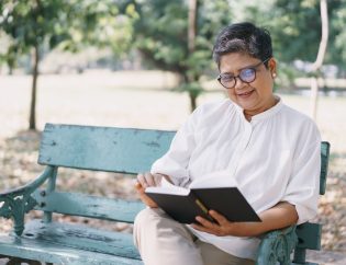 Middle aged Asian woman sitting on a park bench reading a book smiling.