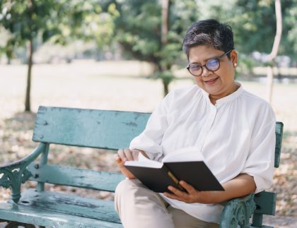 Middle aged Asian woman sitting on a park bench reading a book smiling.