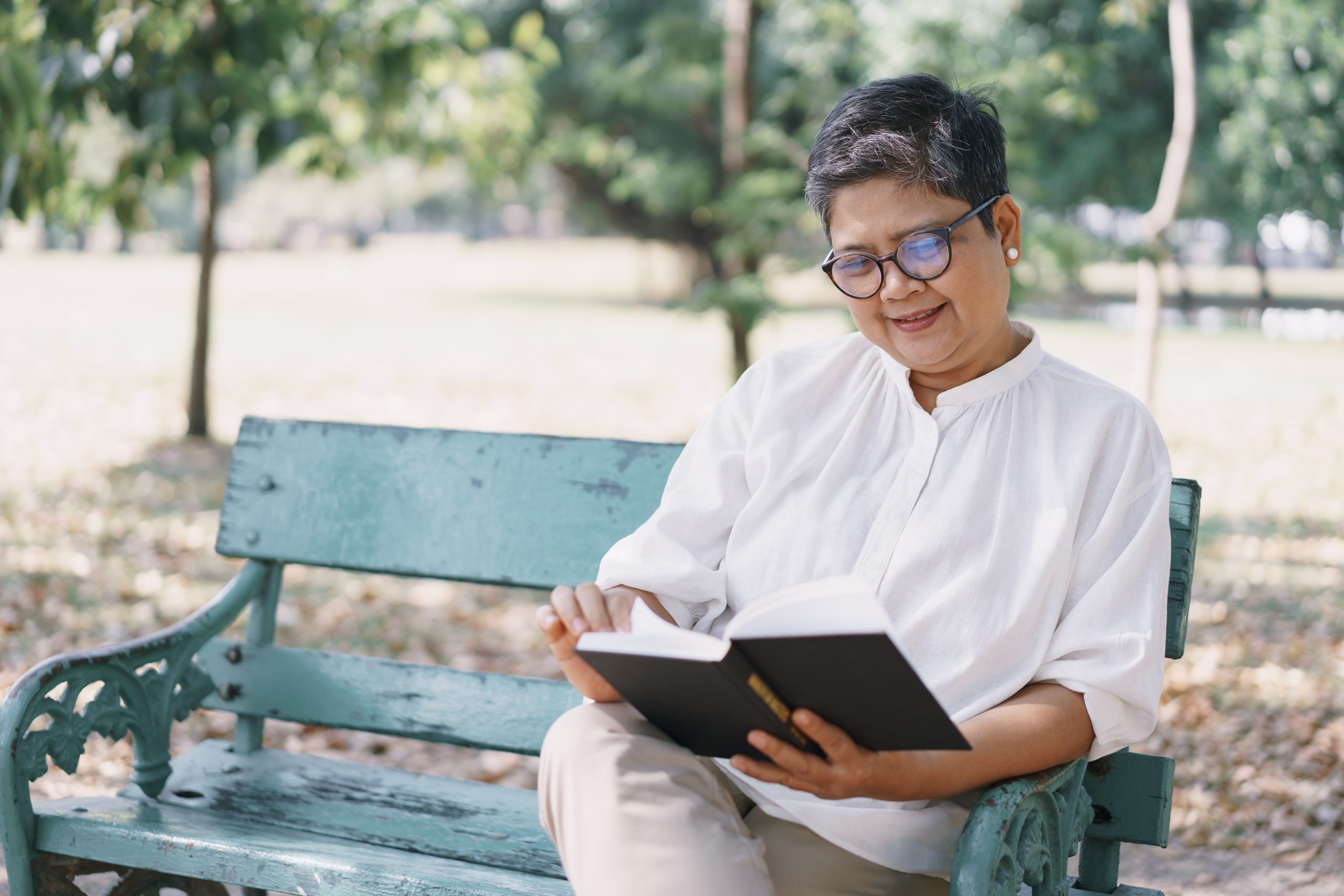 Middle aged Asian woman sitting on a park bench reading a book smiling.