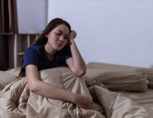 how sleep affects mental health; a young Asian woman struggling to sleep. She is lying awake in her bed with her hand over her head.