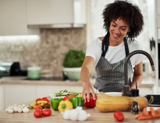 how nutrition and mental health are connected...a young black woman cooking in the kitchen with an array of vegetables scattered around the table in front of her. She is reaching for the red bell pepper.