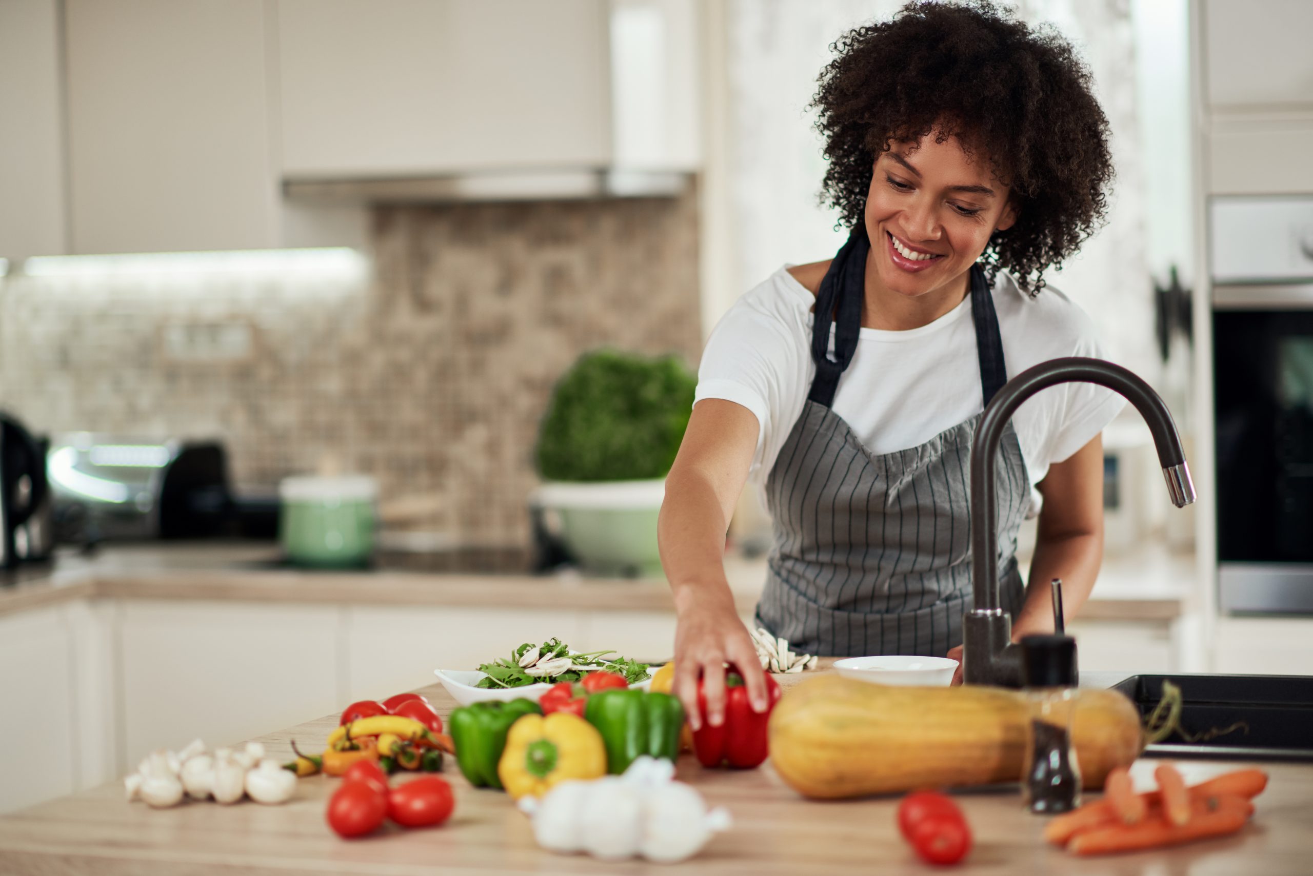 how nutrition and mental health are connected...a young black woman cooking in the kitchen with an array of vegetables scattered around the table in front of her. She is reaching for the red bell pepper.